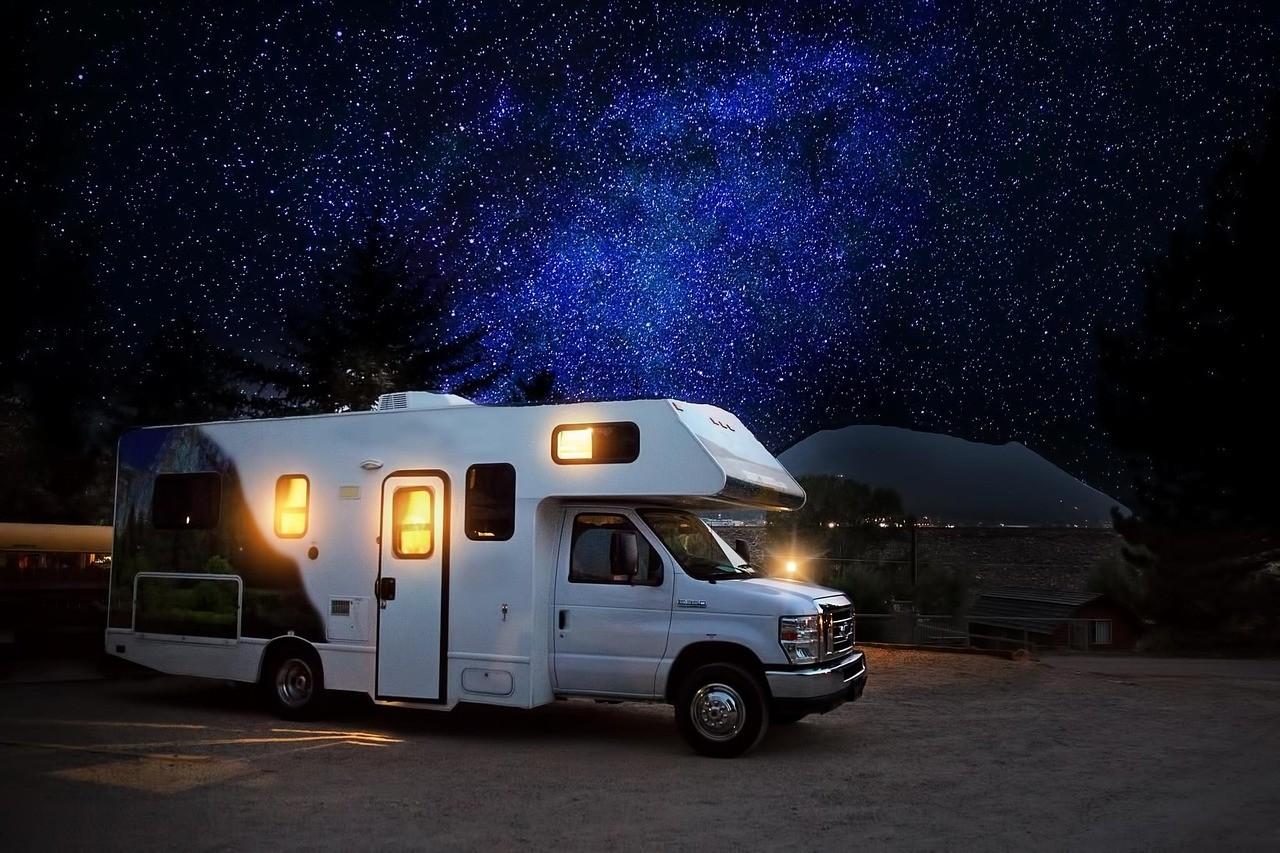An RV parked on open ground at night with its windows lit, beneath a sky full of stars