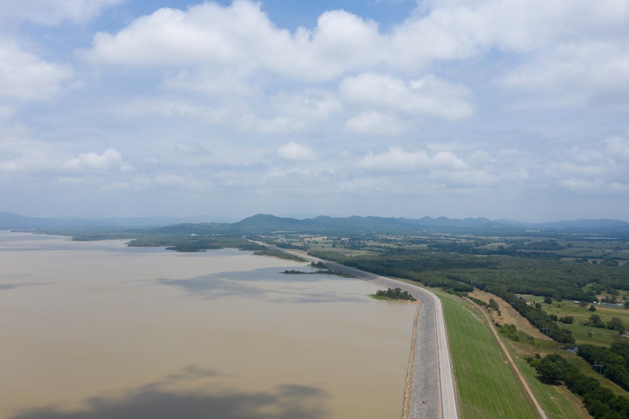 Aerial view of a reservoir in southeastern Oklahoma, its earthen dam and road curving along the shoreline with wooded hills beyond