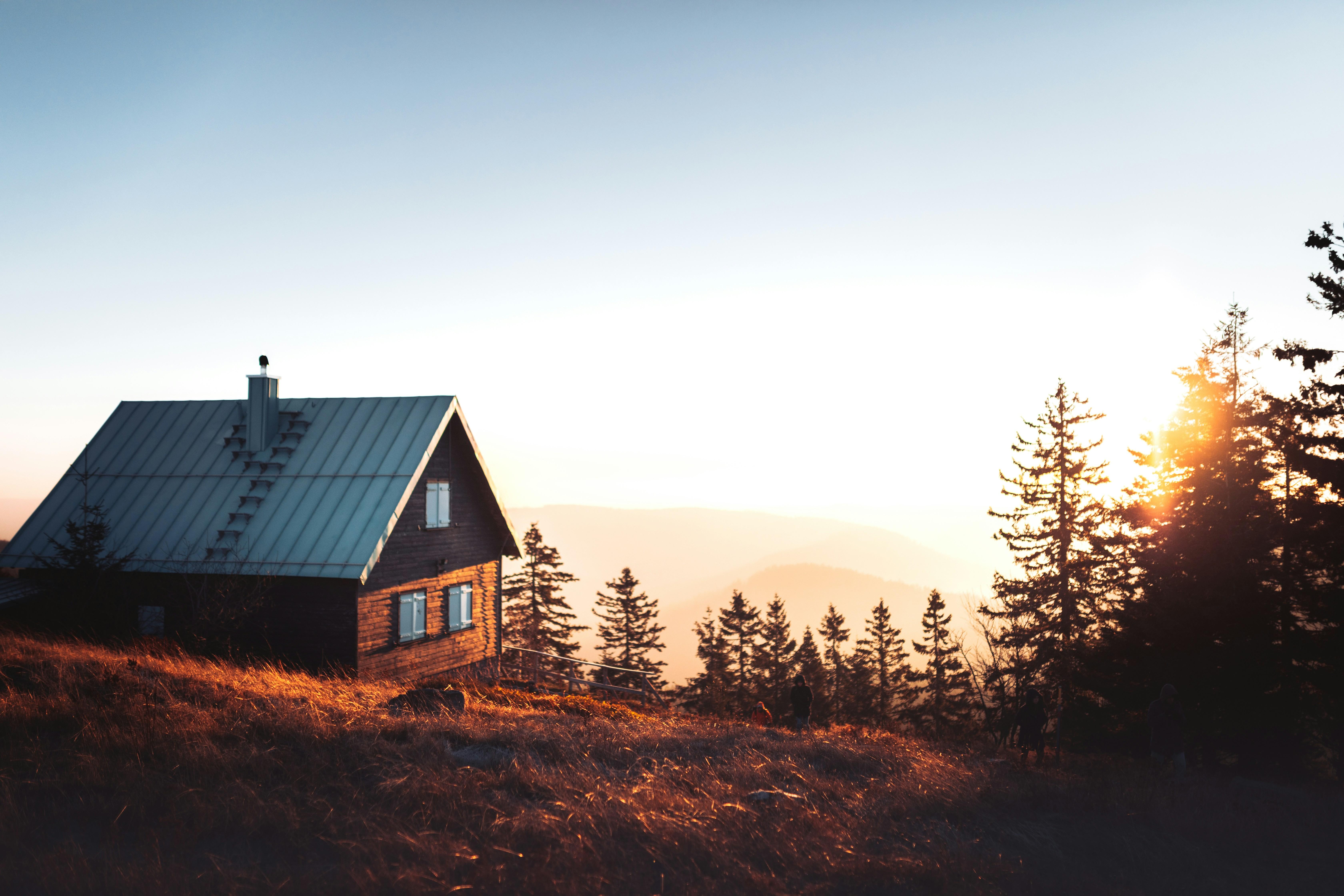 A wooden cabin with a metal roof on a grassy hilltop at sunset, with pines and distant hills behind it